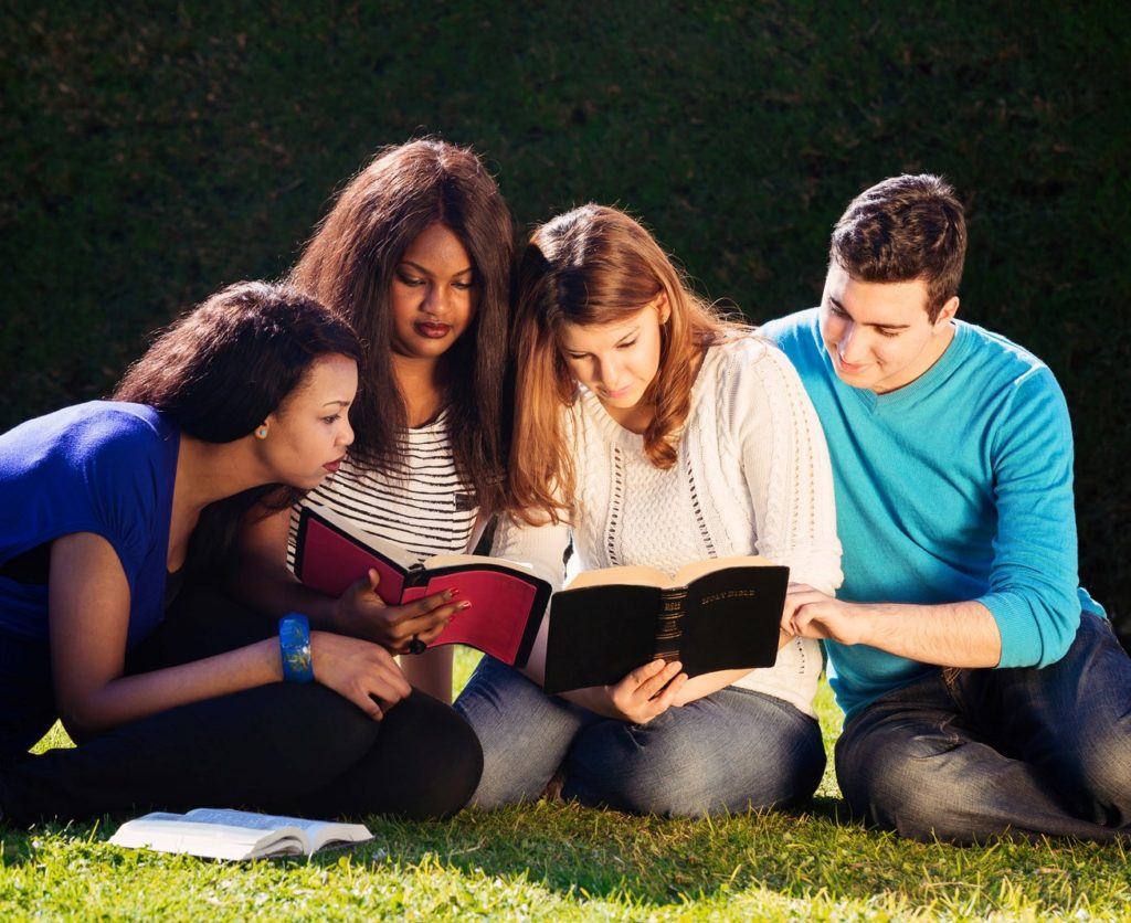 group of one man and two women sitting on the grass reading the Bible together. 