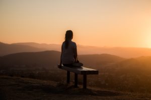 The back of woman sitting on a bench looking at a sunset over rolling hills