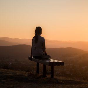 The back of woman sitting on a bench looking at a sunset over rolling hills